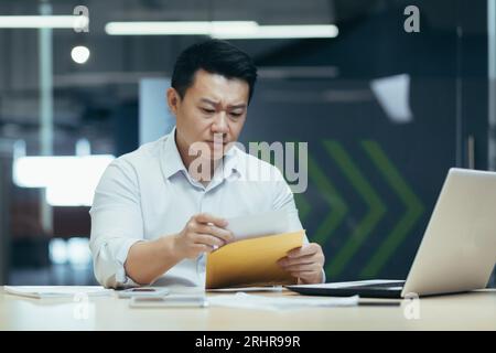 Worried young asian man working in office at laptop and reading received letter, received bills, unpleasant message. Stock Photo