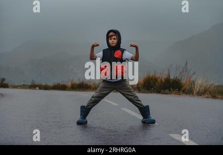 Boy in a heroic pose stands on an asphalt road in rainy weather Stock Photo