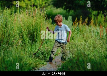Boy in rubber boots with stick walks along a shallow stream Stock Photo