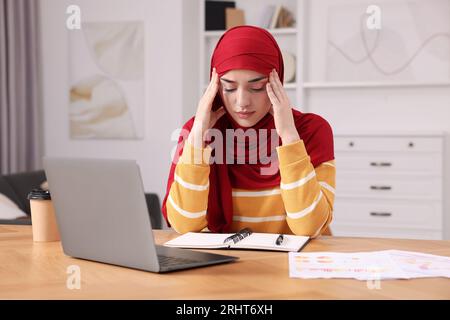 Tired Muslim woman working near laptop at wooden table in room Stock Photo
