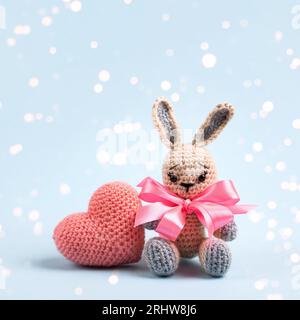 Knitted gray bunny with a crocheted pink heart on a blue background. Creative knitting of toys for children, home decoration. Love concept. Stock Photo