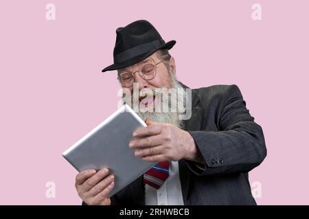 Portrait of laughing aged senior man with tablet pc. Happy grandpa with modern gadget. Isolated n pink. Stock Photo