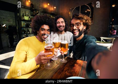Selfie of three male friends toasting happily in a bar, looking at camera with big smiles Stock Photo