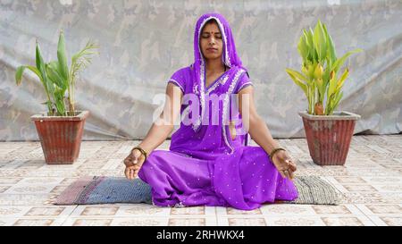 Full length mindful young indian woman making mudra gesture, sitting in lotus position on comfortable couch at home. Peaceful millennial girl deeply m Stock Photo