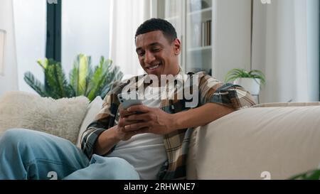 African American guy ethnic happy smiling man relaxing on comfortable couch sofa at home using mobile application scrolling phone app chatting social Stock Photo