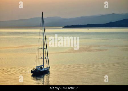 A single yacht sailing on calm waters in the early morning sunrise with distant mountains and hills in the distance Stock Photo