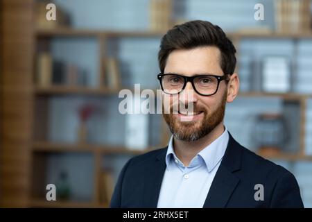 Close-up portrait of mature adult experienced businessman, boss smiling and looking at camera wearing glasses and business suit, successful investor at workplace inside office building. Stock Photo
