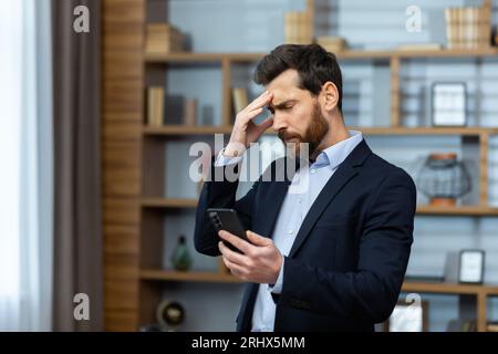 Serious thinking businessman standing near window at workplace, senior boss received online notification on phone with bad news, man in suit reading disappointed, using app on smartphone. Stock Photo