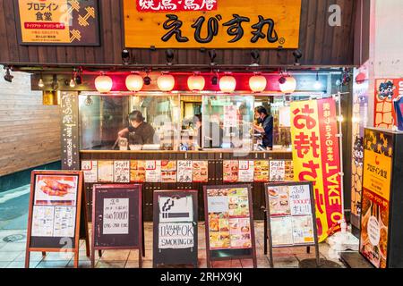 Ebisumaru yakitori restaurant in Shinshigai shopping street, Kumamoto. Extorier, take away counter with three chefs working, diners inside, night. Stock Photo