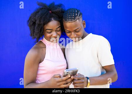 Portrait of positive African American teenage couple with Afro hairstyle browsing mobile phone while standing together on blue background in studio Stock Photo