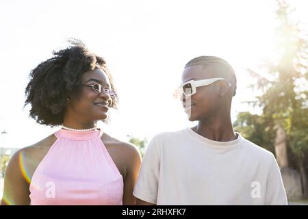 Portrait of young African American teenage couple in trendy outfit and stylish sunglasses smiling and looking at each other in park on sunny day Stock Photo