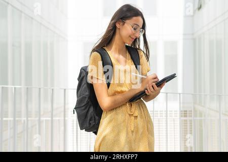 Side view of smiling young woman in yellow dress with long brown hair using digital tablet with pen while standing against blue background Stock Photo
