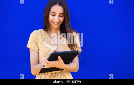 Smiling young woman in yellow dress with long brown hair using digital tablet while standing against blue background Stock Photo