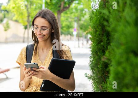 Smiling young female in young dress and eyeglasses browsing smartphone while standing with backpack and tablet against modern building and green bush Stock Photo