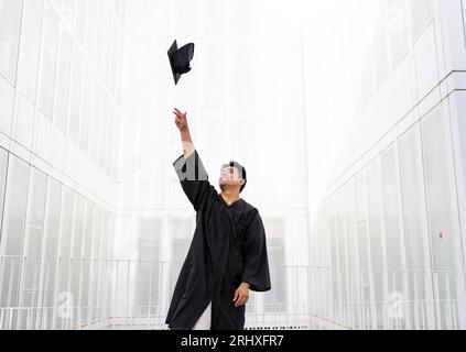 Cheerful Caucasian young male student wearing black graduation gown throwing mortarboard in mid-air while standing against white wall Stock Photo