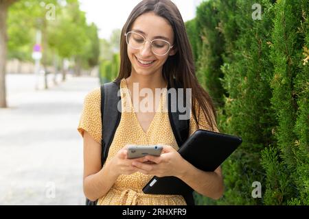 Smiling young female in young dress and eyeglasses browsing smartphone while standing with backpack and tablet against modern building and green bush Stock Photo