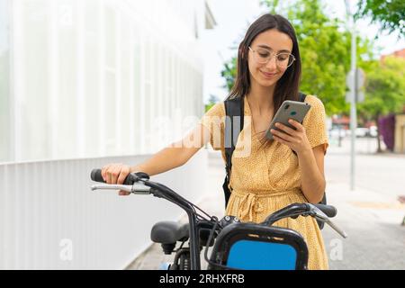 Smiling young female in yellow dress with backpack browsing mobile phone while standing with bicycle near white building Stock Photo