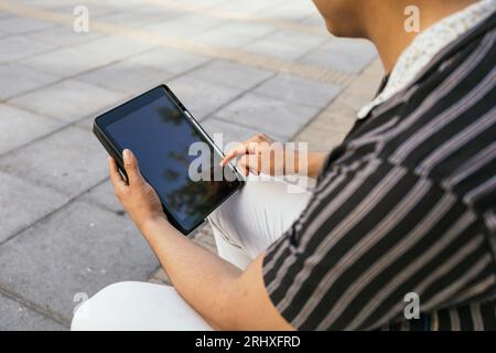 Cropped image of young man dressed in casual outfit holding and using digital tablet while sitting on footpath in city during leisure time Stock Photo