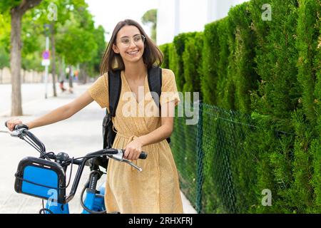 Positive young female in casual clothes smiling and looking away while walking with bicycle near green bushes on street in city Stock Photo