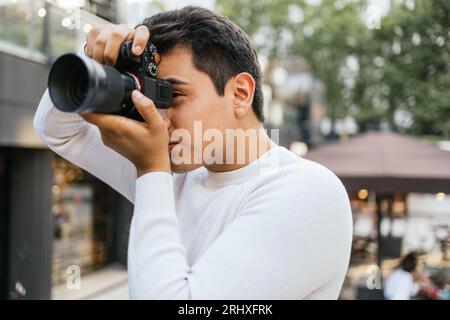 Caucasian young man wearing white t-shirt photographing with camera while standing against blurred background in city Stock Photo