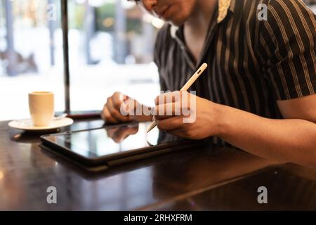 Anonymous handsome young male designer drawing on graphics tablet with digitized pen and having coffee while sitting by window in cafes Stock Photo