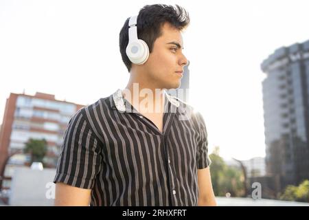 From below Caucasian thoughtful handsome young man listening to music over earphones and looking away seriously while standing against clear sky in ci Stock Photo