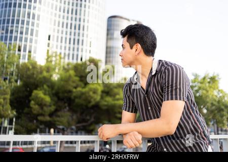 Caucasian handsome young man wearing black shirt contemplating and looking away while leaning on railing against buildings and clear sky in city Stock Photo