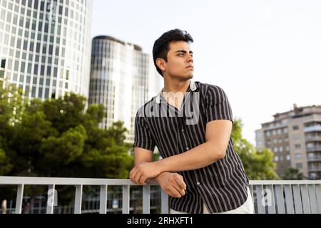 Caucasian handsome young man wearing black shirt contemplating and looking away while leaning on railing against buildings and clear sky in city Stock Photo