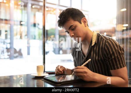 Caucasian handsome young male designer drawing on graphics tablet with digitized pen and having coffee while sitting by window in cafes Stock Photo