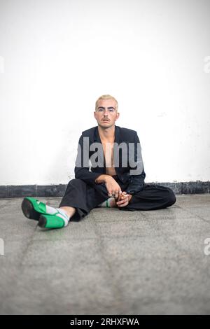 Confident male in high heels looking at camera while sitting alone with stretched leg on marble floor of empty room against white wall in daylight Stock Photo