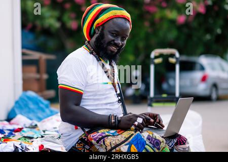 Side view of African American man in colorful clothing and hat sitting and looking at camera while typing on laptop against blurred background in dayl Stock Photo