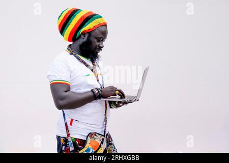 Side view of African American man in colorful clothing and hat standing and looking at screen while typing on laptop against white background Stock Photo