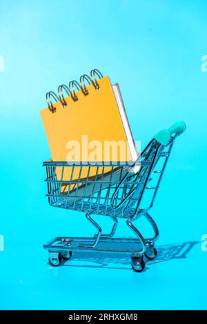 Top spiral bound yellow hardcovers notebook placed in miniature supermarket shopping trolley with shadow in light against vivid blue background Stock Photo
