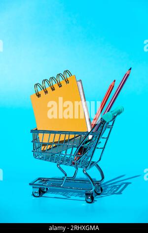 Top spiral bound yellow hardcovers notebook and colorful pencils placed in miniature supermarket shopping trolley with shadow in light against vivid b Stock Photo