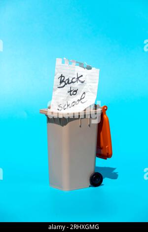 Holes on top torn paper with pencil message back to school placed in miniature gray dust bin with red top cover black wheel and shadow in light agains Stock Photo