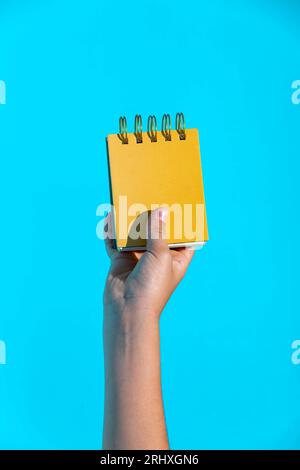 Crop hand of anonymous person holding small yellow spiral notepad against bright blue background Stock Photo