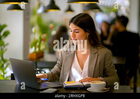 Concentrated young female freelancer in formal cloth sitting at cafe table with laptop and cup of coffee with notebook while working on remote project Stock Photo