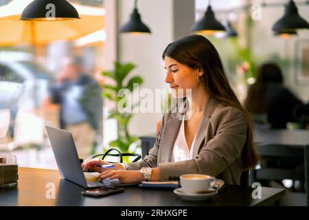 Concentrated young female freelancer in formal cloth sitting at cafe table with laptop and cup of coffee with notebook while working on remote project Stock Photo