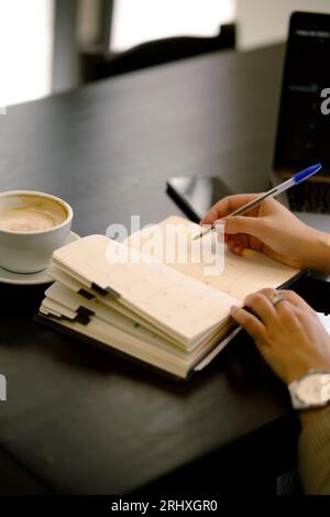 Anonymous young while sitting at table with notebook and taking notes with pen in cozy blurred cafe Stock Photo