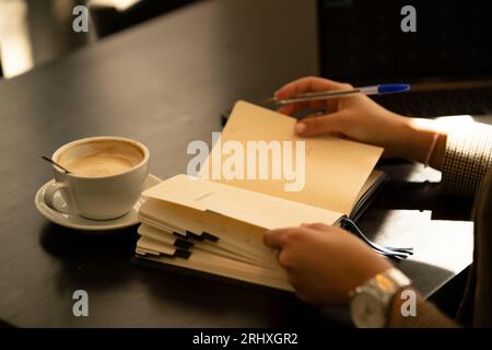 Anonymous young while sitting at table with notebook and taking notes with pen in cozy blurred cafe Stock Photo