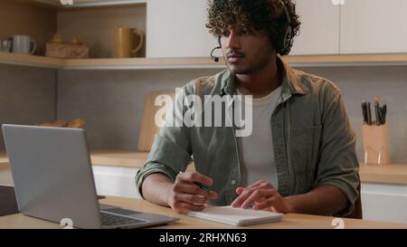 Home education at quarantine self-education Arabian man in headset listen webinar class lesson studying online in kitchen writing watching laptop Stock Photo