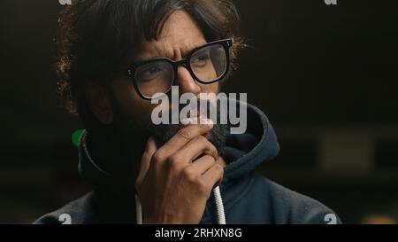 Close up Indian pensive man in eyeglasses thinking looking to side rubbing chin. Thoughtful Arabian male in glasses stand at dark parking lot Stock Photo