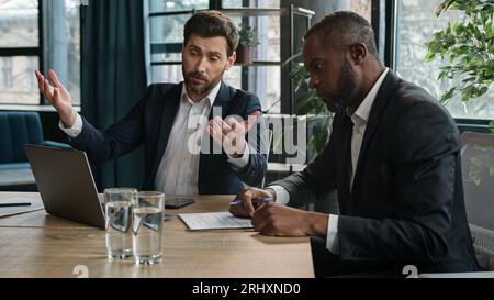 Caucasian manager showing African boss bad business result computer mistake two diverse men multiracial coworkers businessmen with papers and laptop Stock Photo