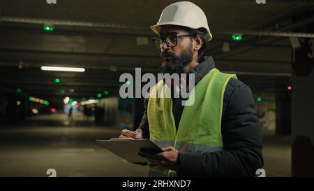 Indian contractor mechanic in helmet and glasses writing in paper examination parking lot write in clipboard. Arabian bearded man worker of Stock Photo