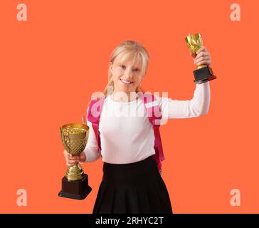 Smart little schoolgirl in stylish uniform with prize cups on orange background Stock Photo