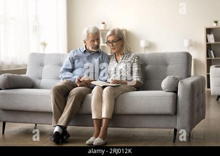 Focused mature wife in glasses reading book to senior husband Stock Photo