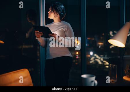 Thoughtful businesswoman using a digital tablet in her office at night, working overtime. She stands next to a window, focused and dedicated to her wo Stock Photo