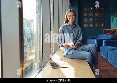 Smiling young woman freelancer sitting near window and making notes during working on laptop Stock Photo