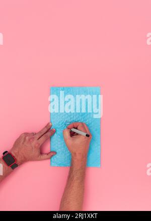 Overhead view of Caucasian adult male hands writing a shipping address on a blue bubble wrap packing envelope. Pink background with copy space. Stock Photo
