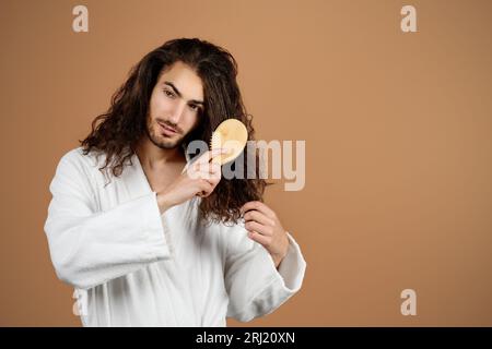 Young Hispanic Guy Brushing Long Curly Hair With Hairbrush, Studio Stock Photo